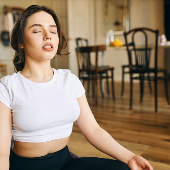 Beautiful young Caucasian woman with muscular curvy body sitting in lotus posture at home keeping eyes closed, meditating during yoga practive, doing body scanning, concentrating on breathing